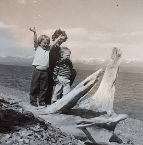 Maria, mother and bother near the shoreline with mountains far off in distance, driftwood in foreground, Maria's arms in the air, happy, smiling