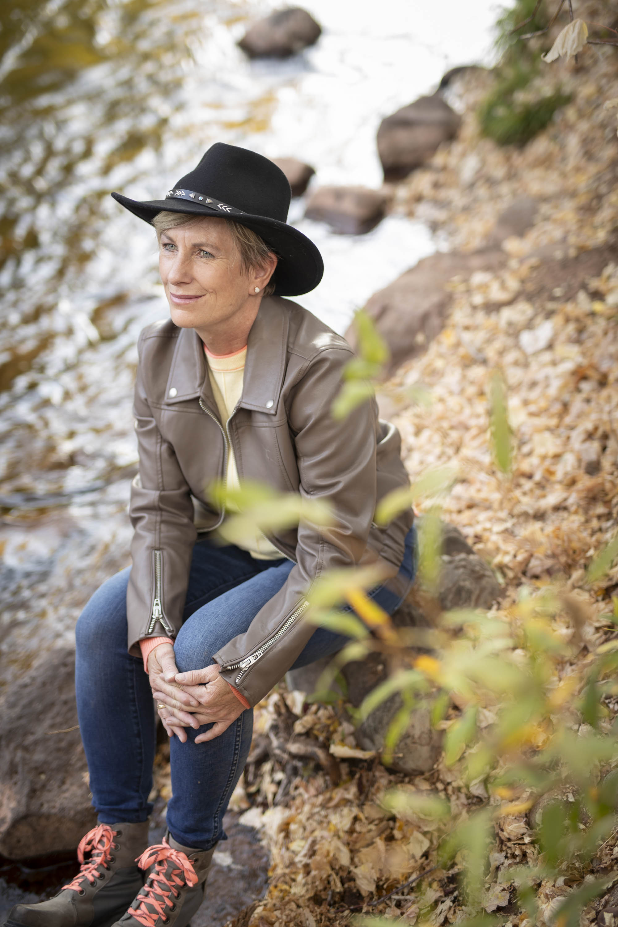 Maria Armstrong - black cowboy Hat, brown leather jacket sitting by a river